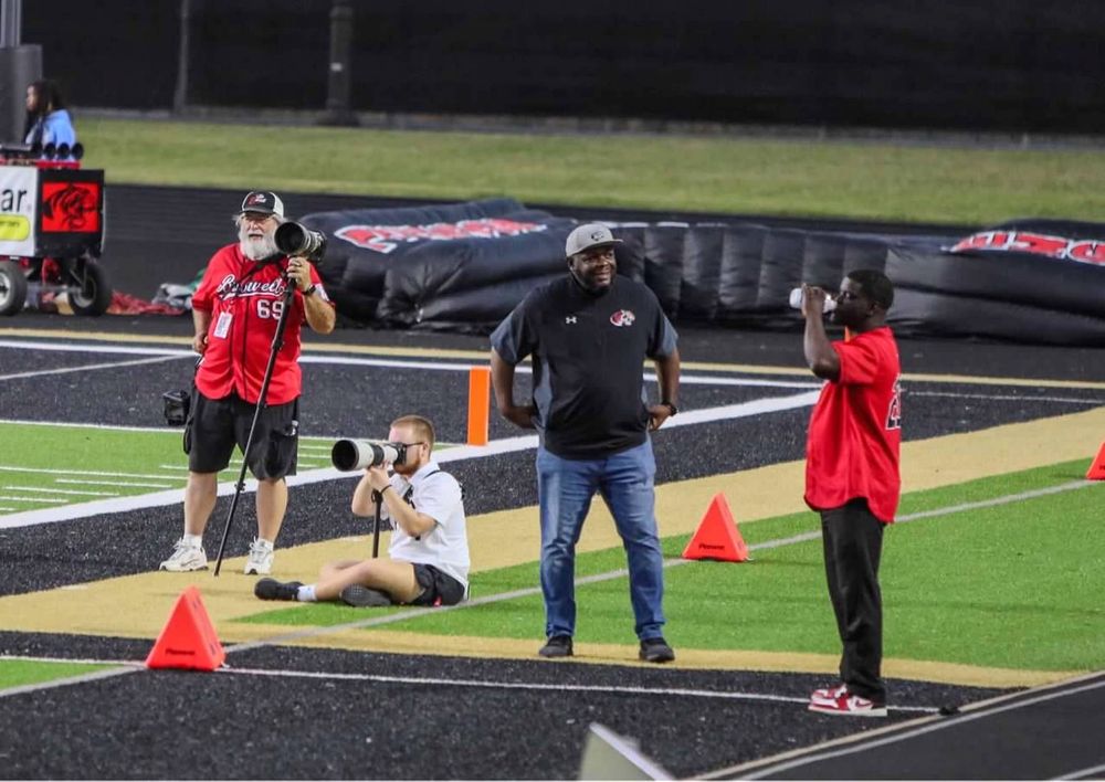 Three sports photographers capture action on football field sideline near orange cones at night.