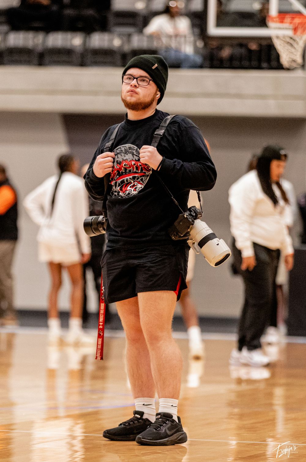 Young male sports photographer stands on basketball court holding camera equipment.