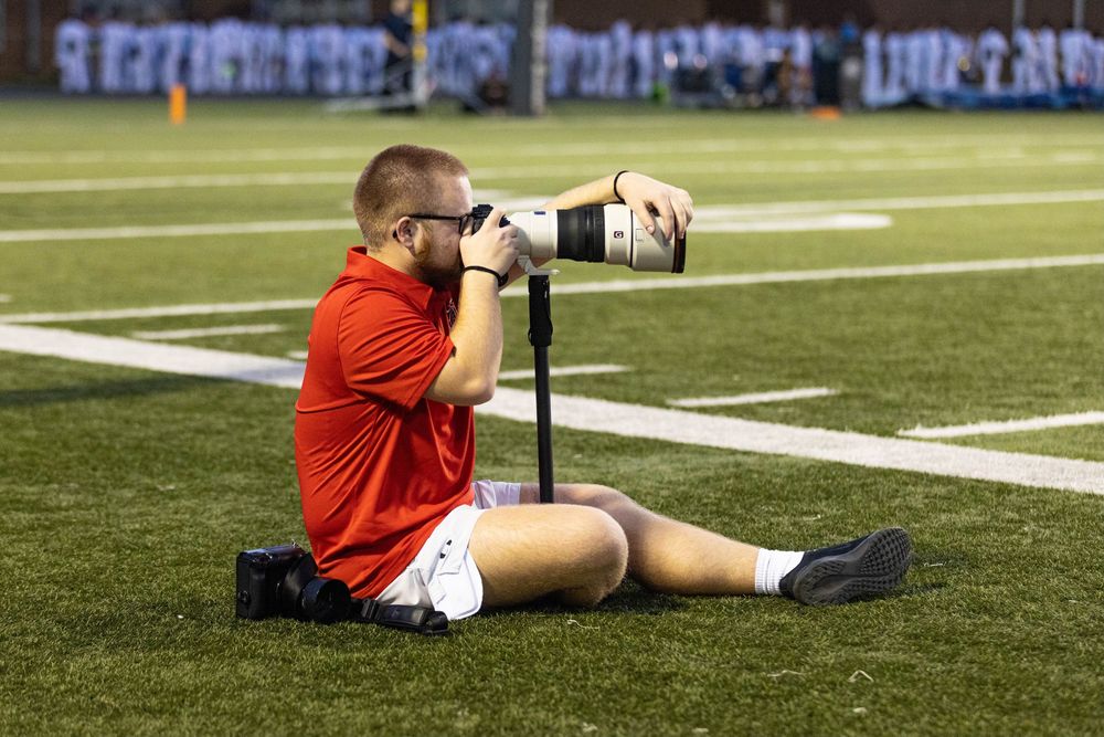 Sports photographer sits on football field using large telephoto lens on monopod.