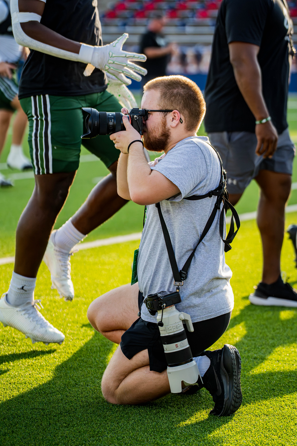 Bearded sports photographer kneels on turf capturing close-up shots during a football game.