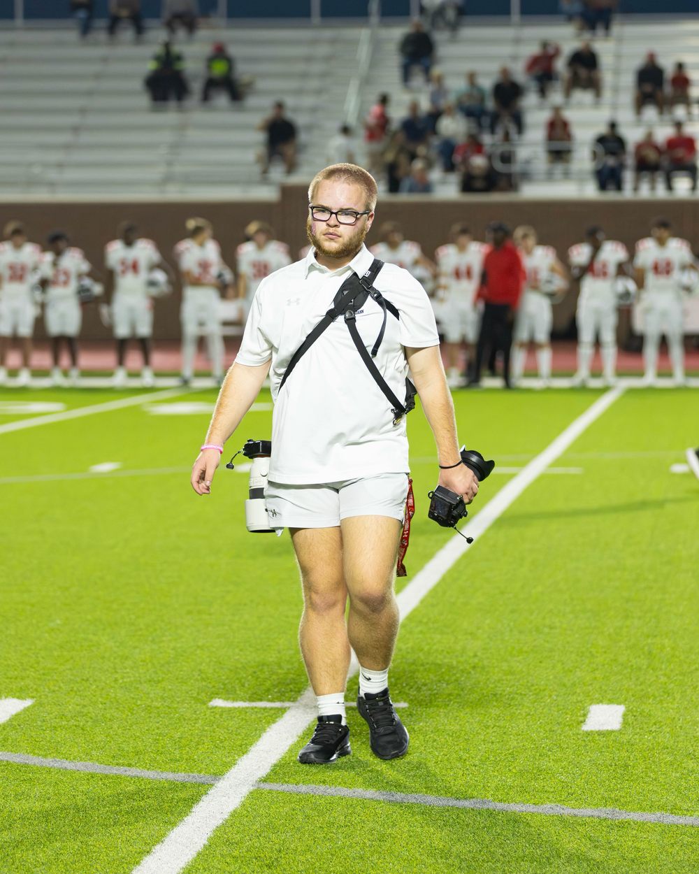 Young photographer carrying dual cameras walks across green football field during a high school game.
