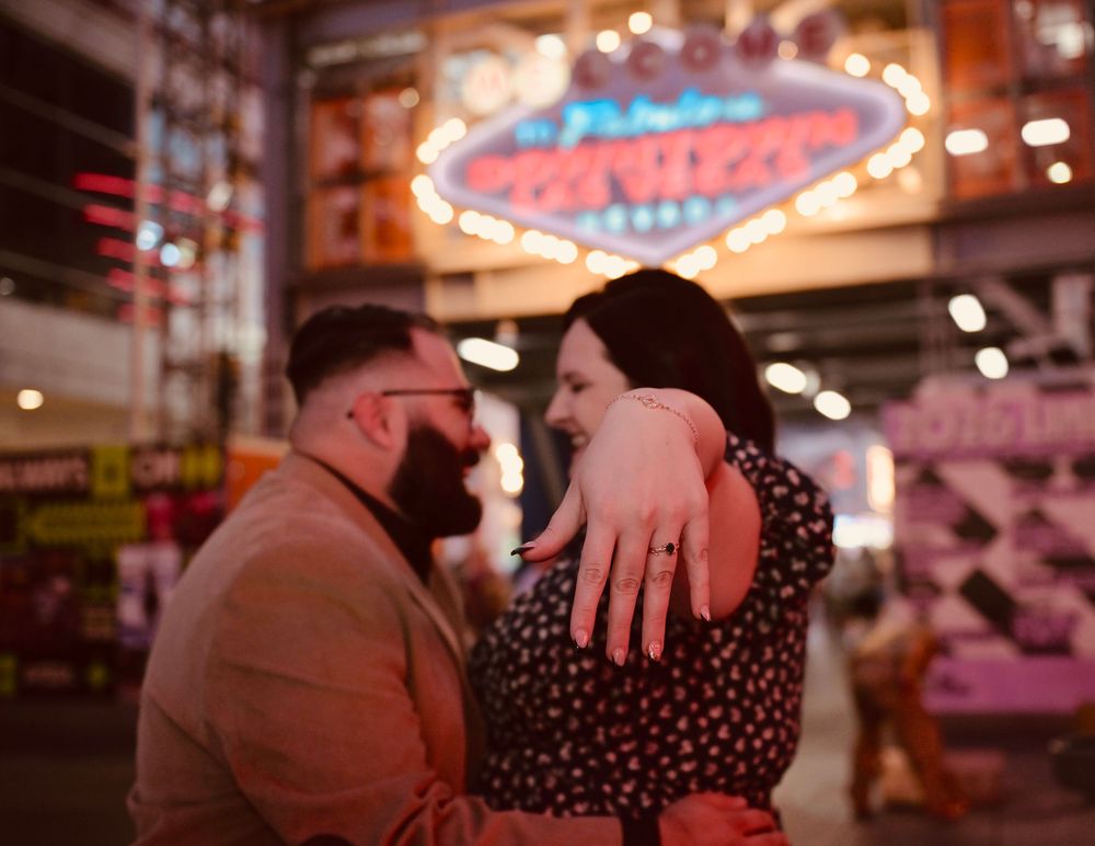 Couple embracing and showing engagement ring near Las Vegas neon signs at night.