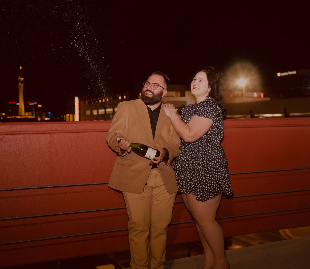 Couple celebrating at night, man popping champagne bottle while woman laughs outdoors.