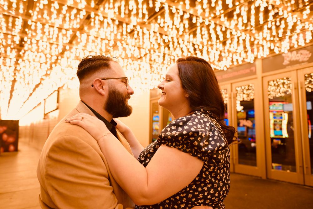 Couple sharing a romantic moment under warm string lights in a glowing venue, woman in floral dress smiling at bearded man.