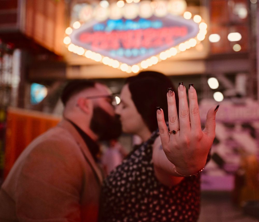 Couple kissing in Las Vegas with engagement ring displayed in foreground under bright neon lights.