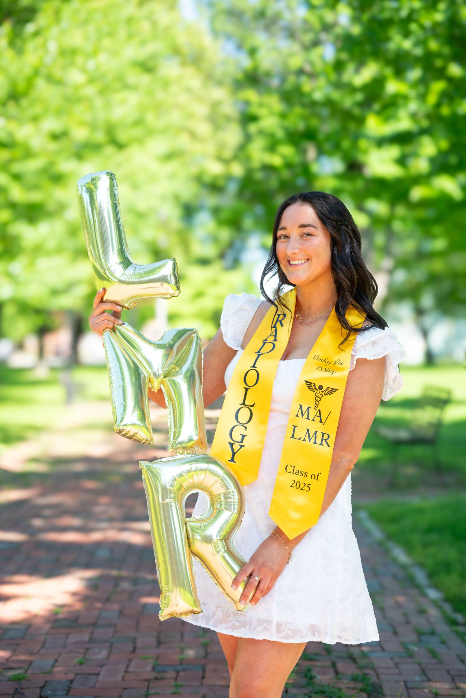 A smiling graduate holds gold letter balloons wearing a yellow Radiology MA/LMR Class of 2025 stole outdoors.