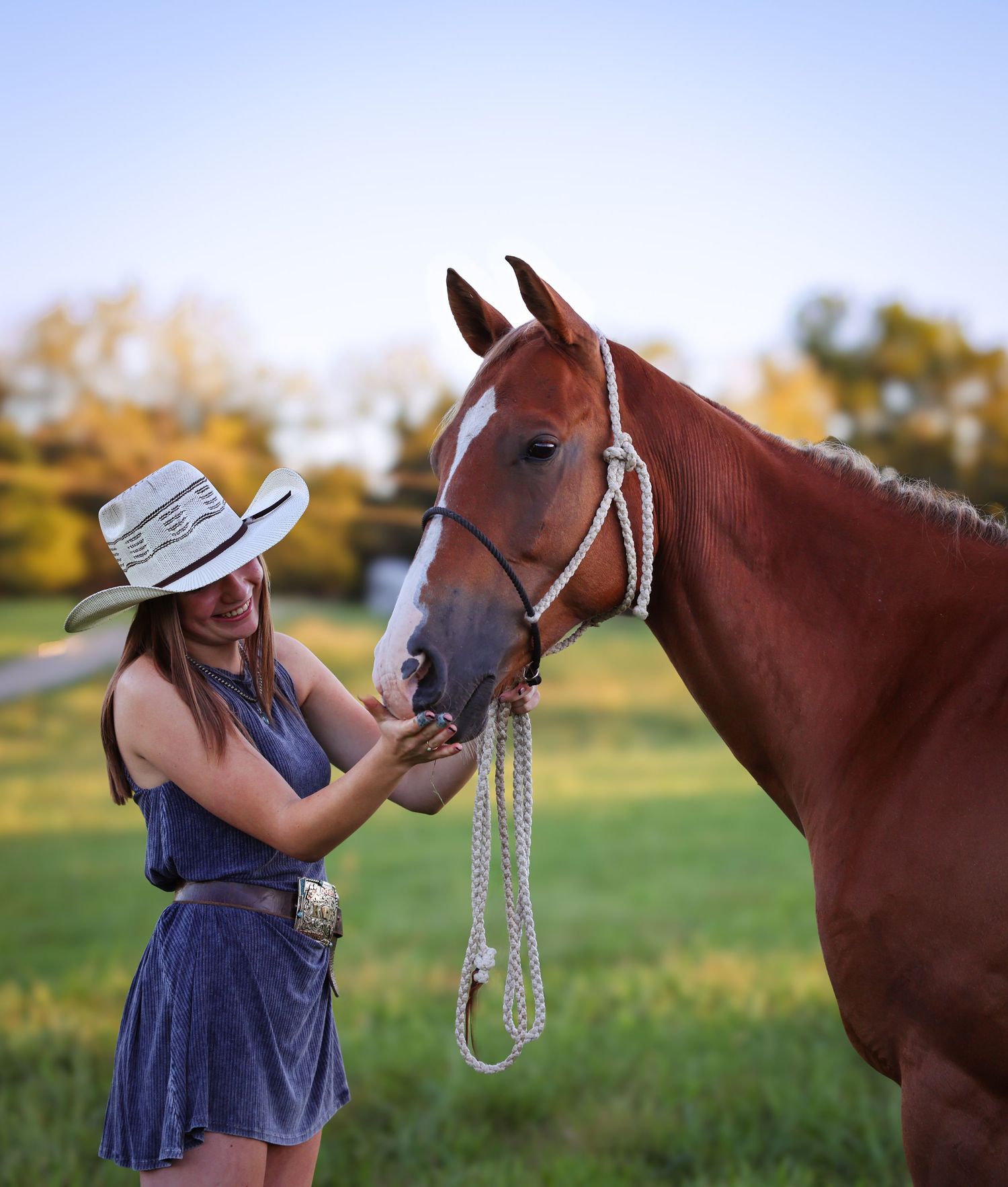 A smiling young woman in a cowboy hat affectionately pets a chestnut horse in a sunny field.