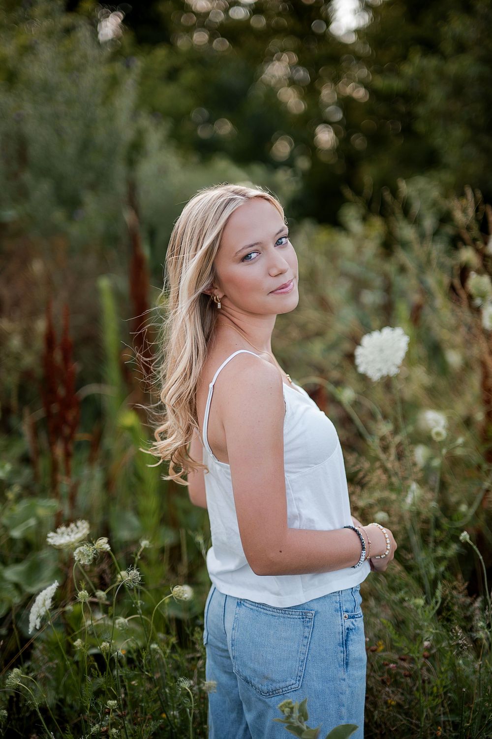 Young blonde woman in white tank top and jeans posing outdoors among wildflowers and greenery.