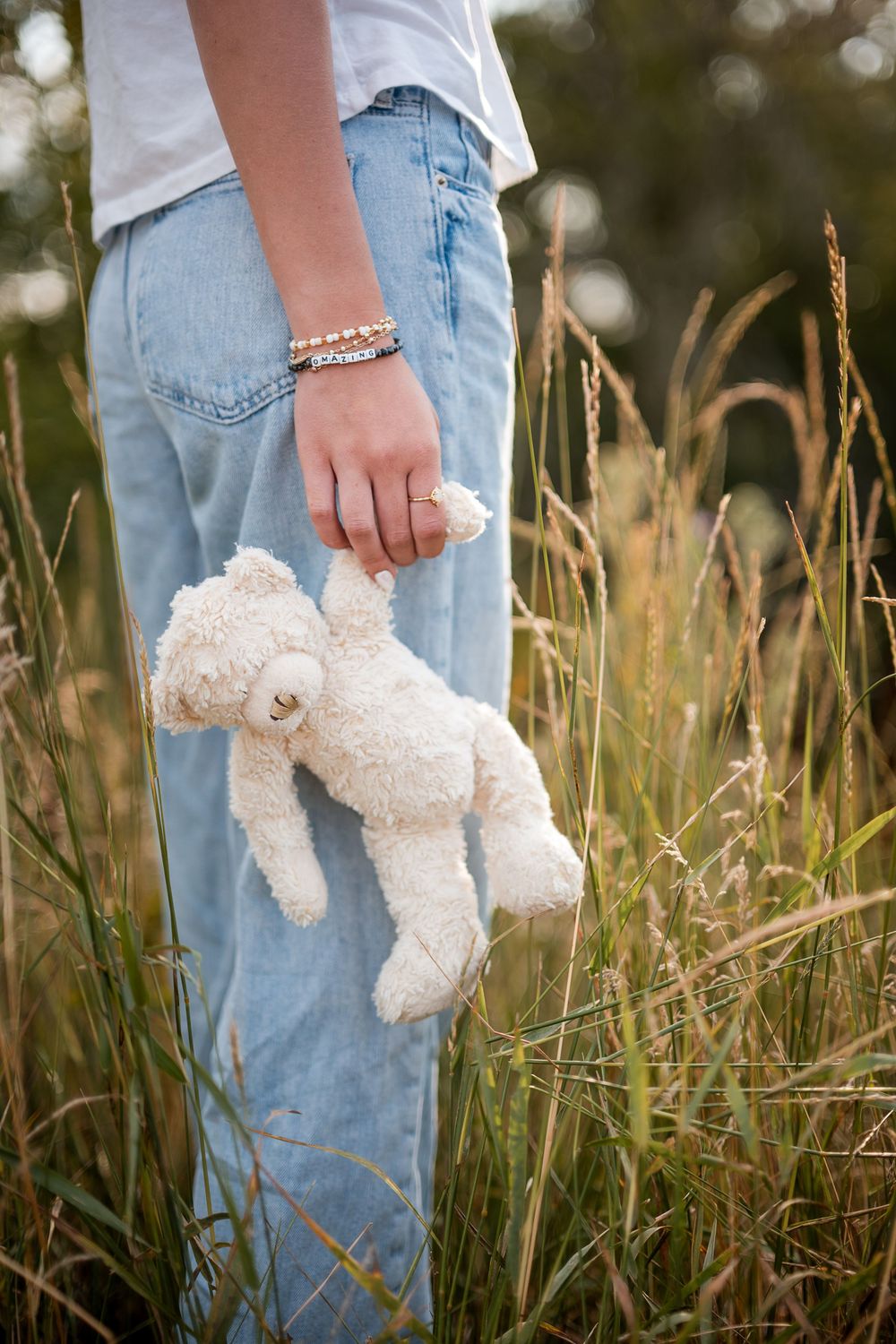 Teen holding a white teddy bear in tall golden grass field, wearing light blue jeans and bracelets.