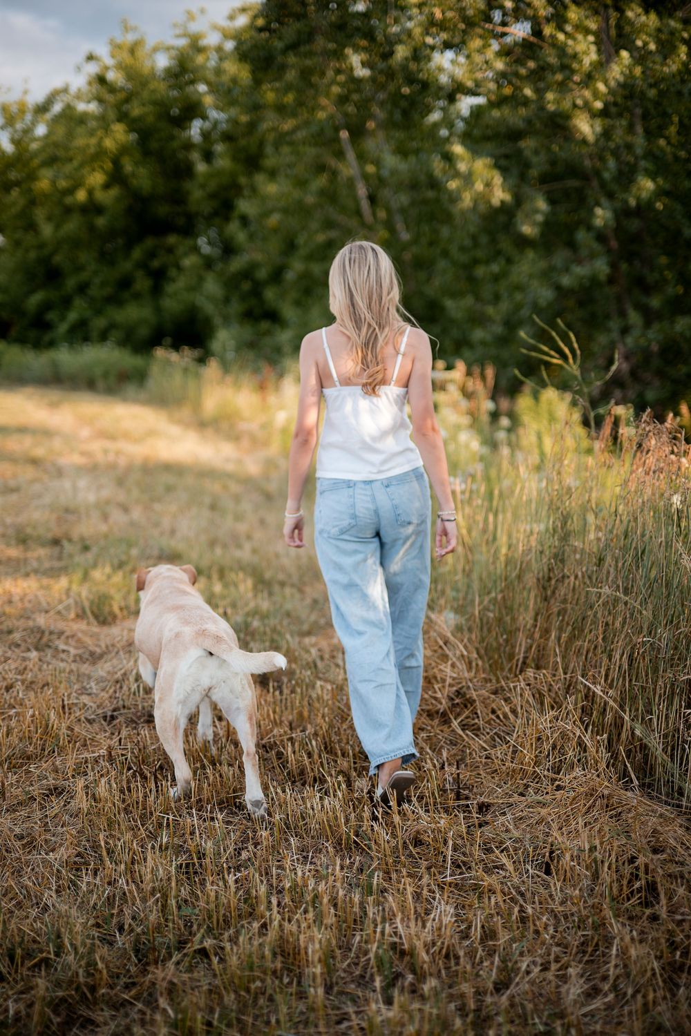 Blonde woman in white top and jeans walking with a golden Labrador through a sunny rural field.