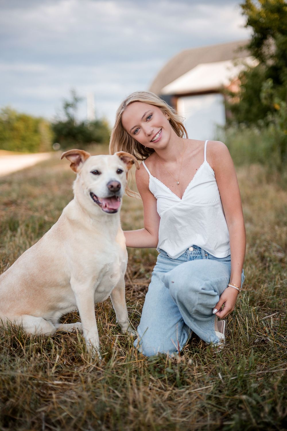 Smiling blonde woman in white top and jeans kneeling beside a happy yellow Labrador mix on a grassy field.