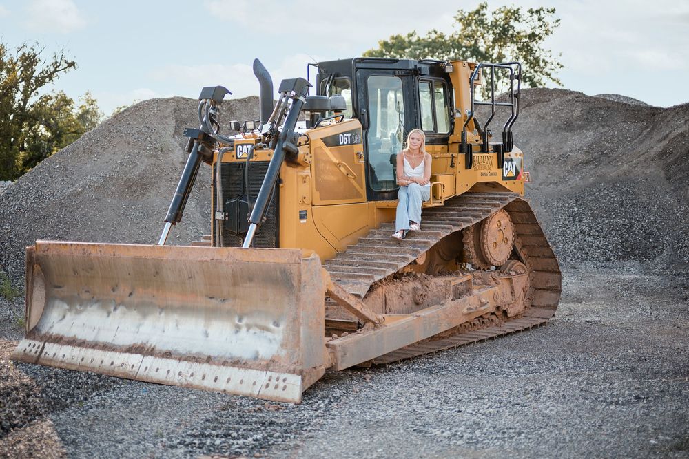 Woman standing beside a large yellow CAT bulldozer on a gravel construction site with dirt mounds in the background.