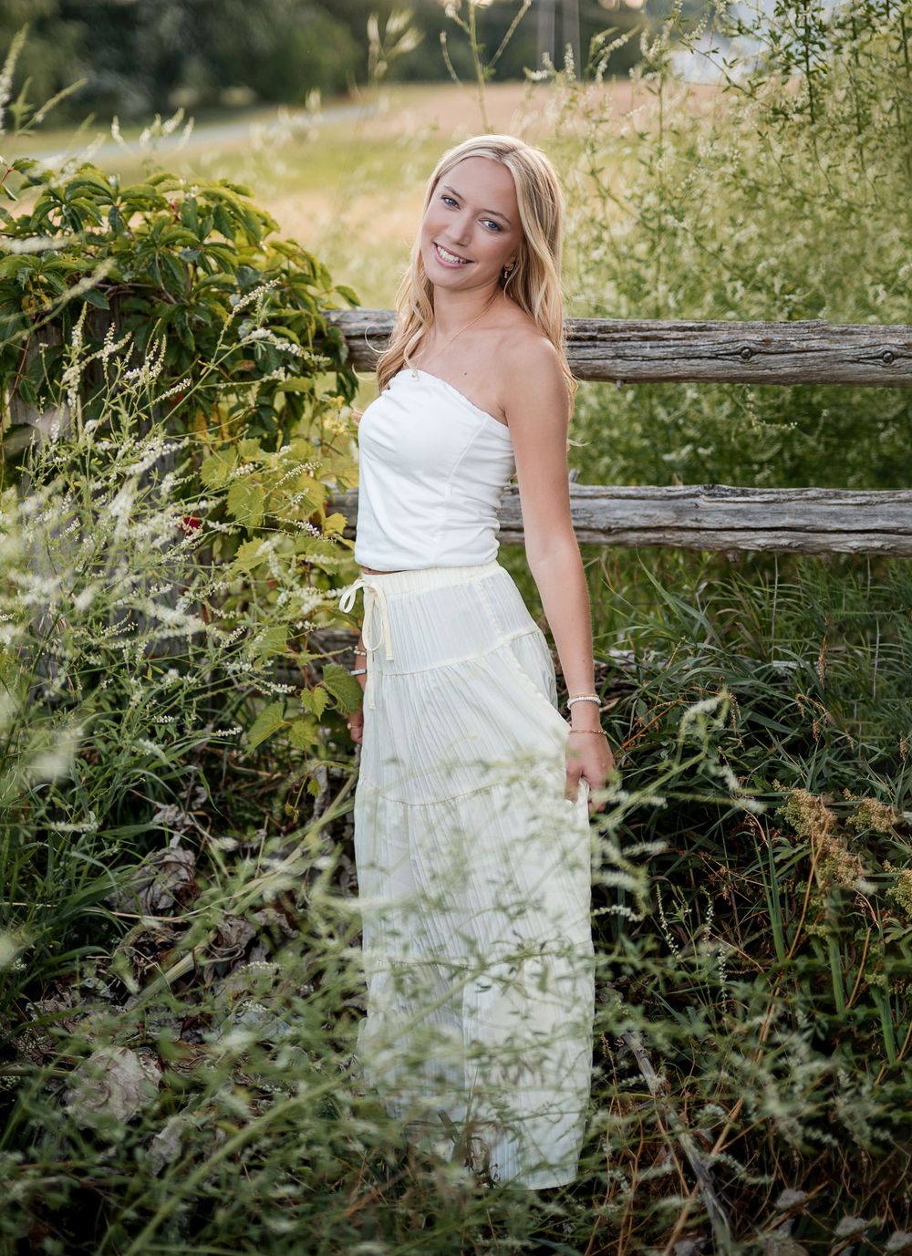 Young blonde woman in white strapless maxi dress smiling outdoors near rustic wooden fence surrounded by lush greenery.