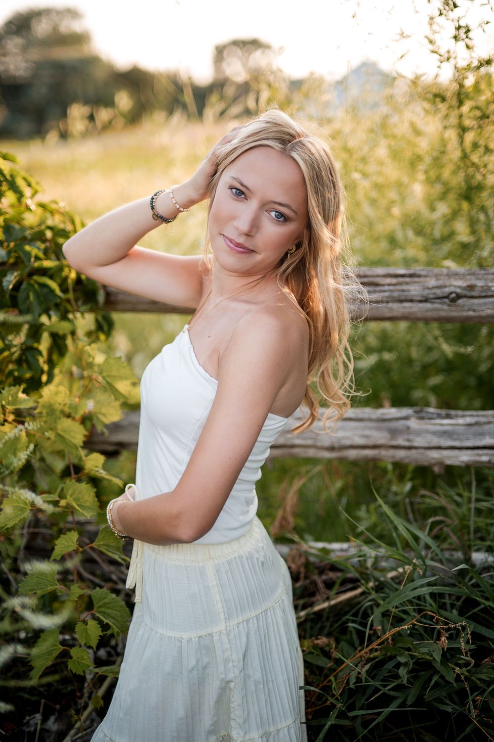 Young blonde woman in white sundress poses gracefully by rustic wooden fence in golden sunlit meadow.