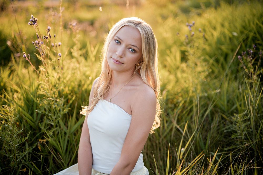 Blonde woman in white strapless dress sitting in golden sunlit green meadow field at sunset.