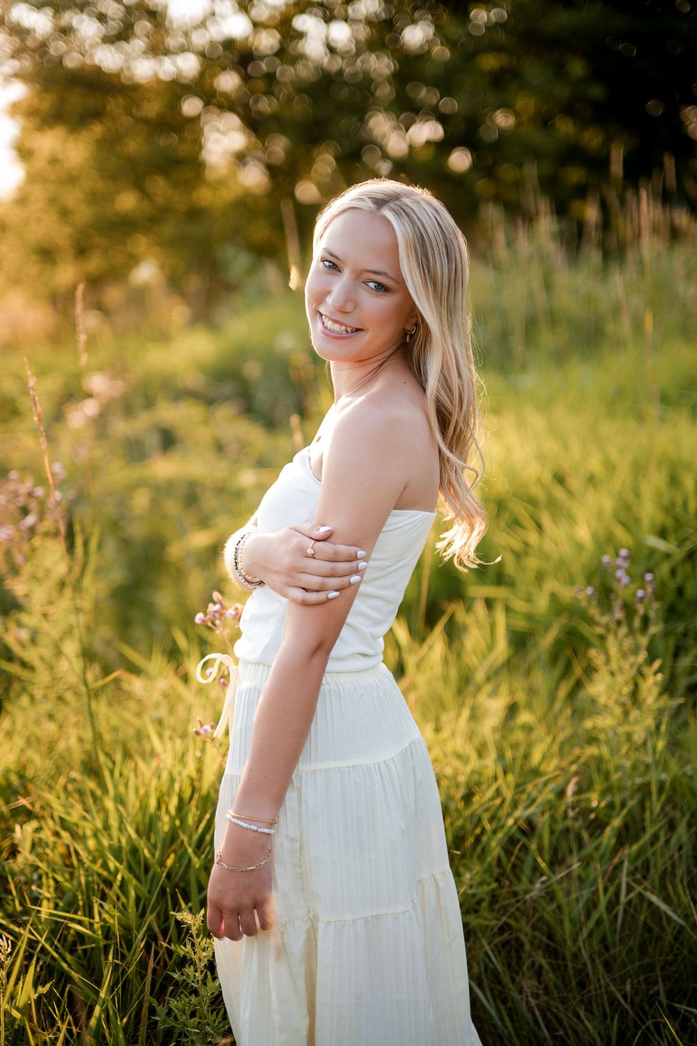 Smiling blonde woman in white sundress standing in golden sunlit meadow with wildflowers at sunset.
