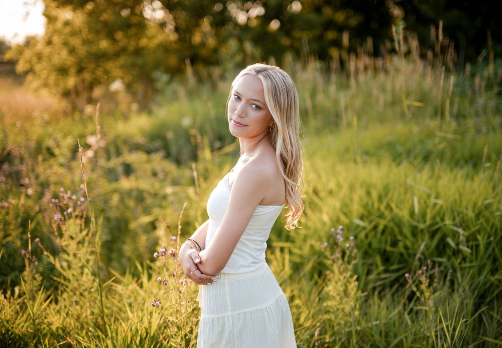 Young blonde woman in white strapless dress posing in sunlit green meadow during golden hour.