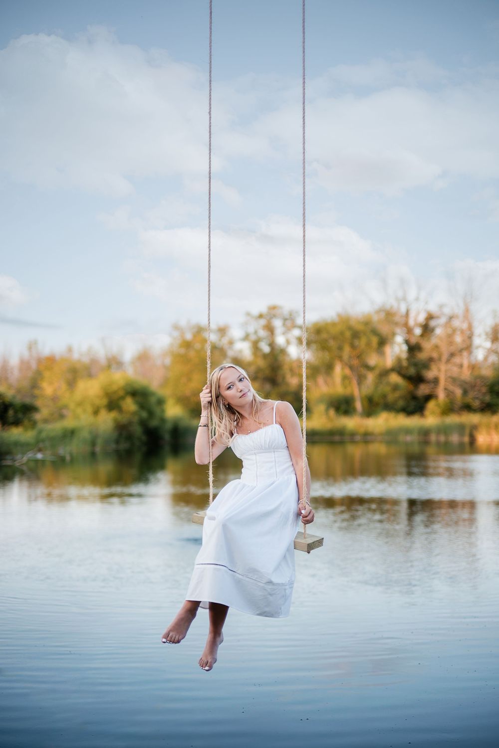 Blonde woman in white dress sitting on a rope swing over a calm lake with green trees in background.