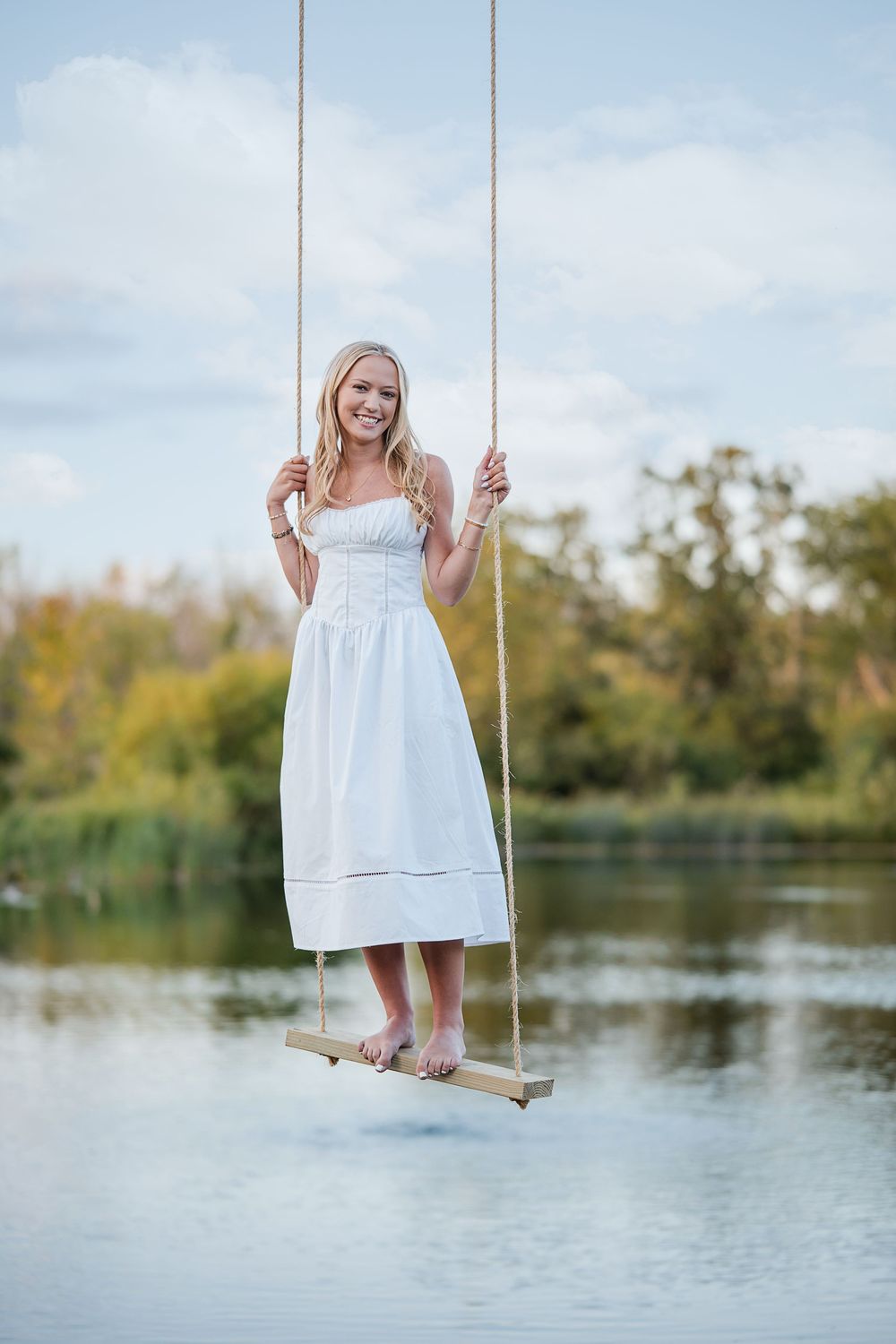 Young blonde woman in white sundress standing barefoot on rope swing over lake with trees in background.