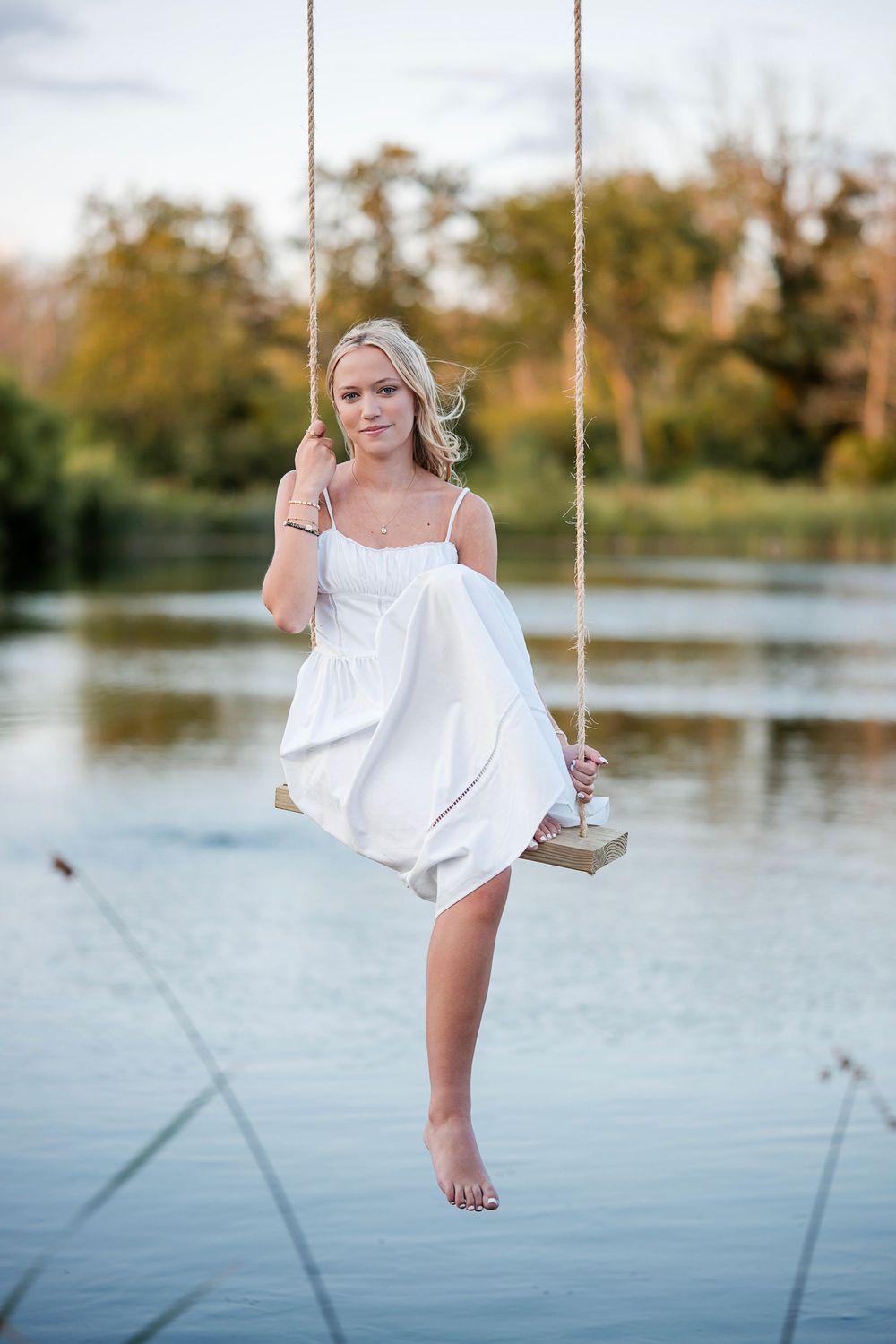 Young blonde woman in white dress sitting on rope swing over calm lake with autumn trees in background.