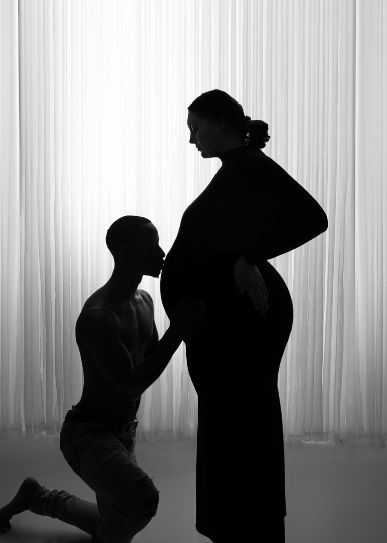Black and white silhouette of a man kneeling and kissing a pregnant woman's belly against a white curtain backdrop.