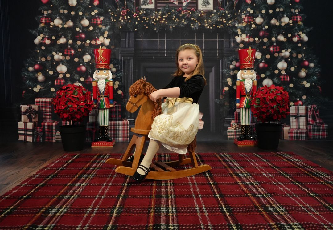 Young girl in holiday dress rides a rocking horse surrounded by Christmas trees, nutcrackers, and poinsettias.