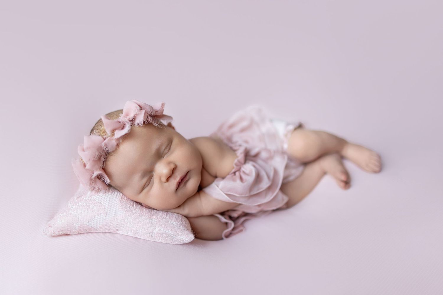 Sleeping newborn baby girl in pink floral headband and ruffle outfit resting on soft pink pillow.