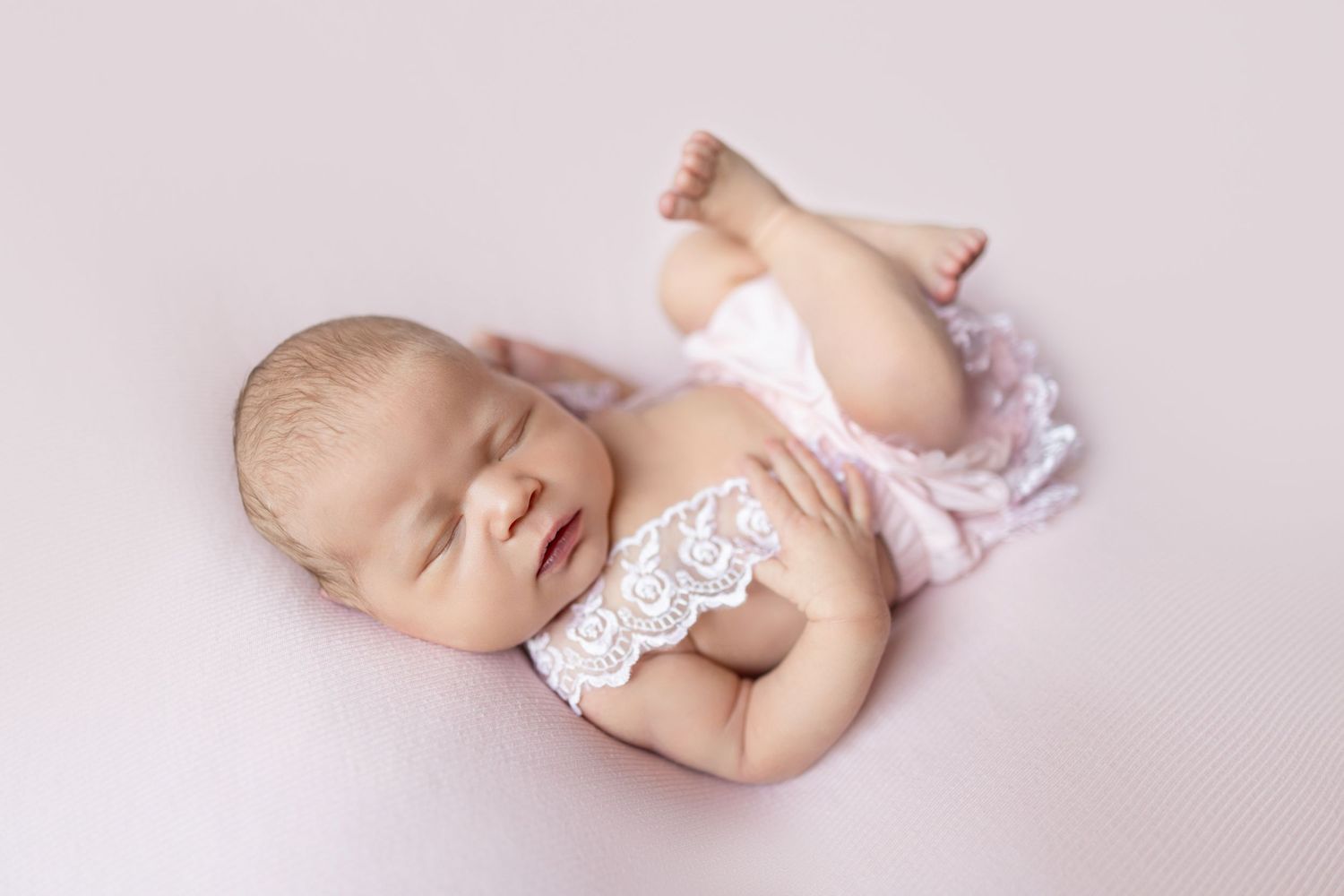 Sleeping newborn baby girl in pink lace romper posed on soft pink backdrop for professional newborn photography session.