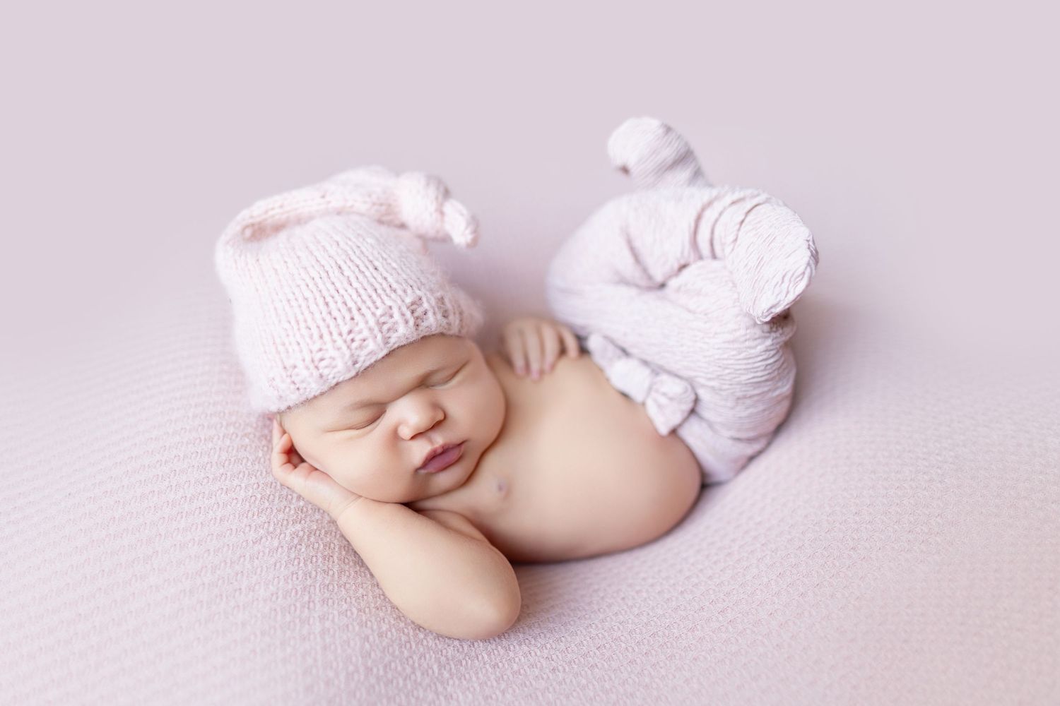 Sleeping newborn baby in pink knit hat and pants curled up on soft lavender blanket.