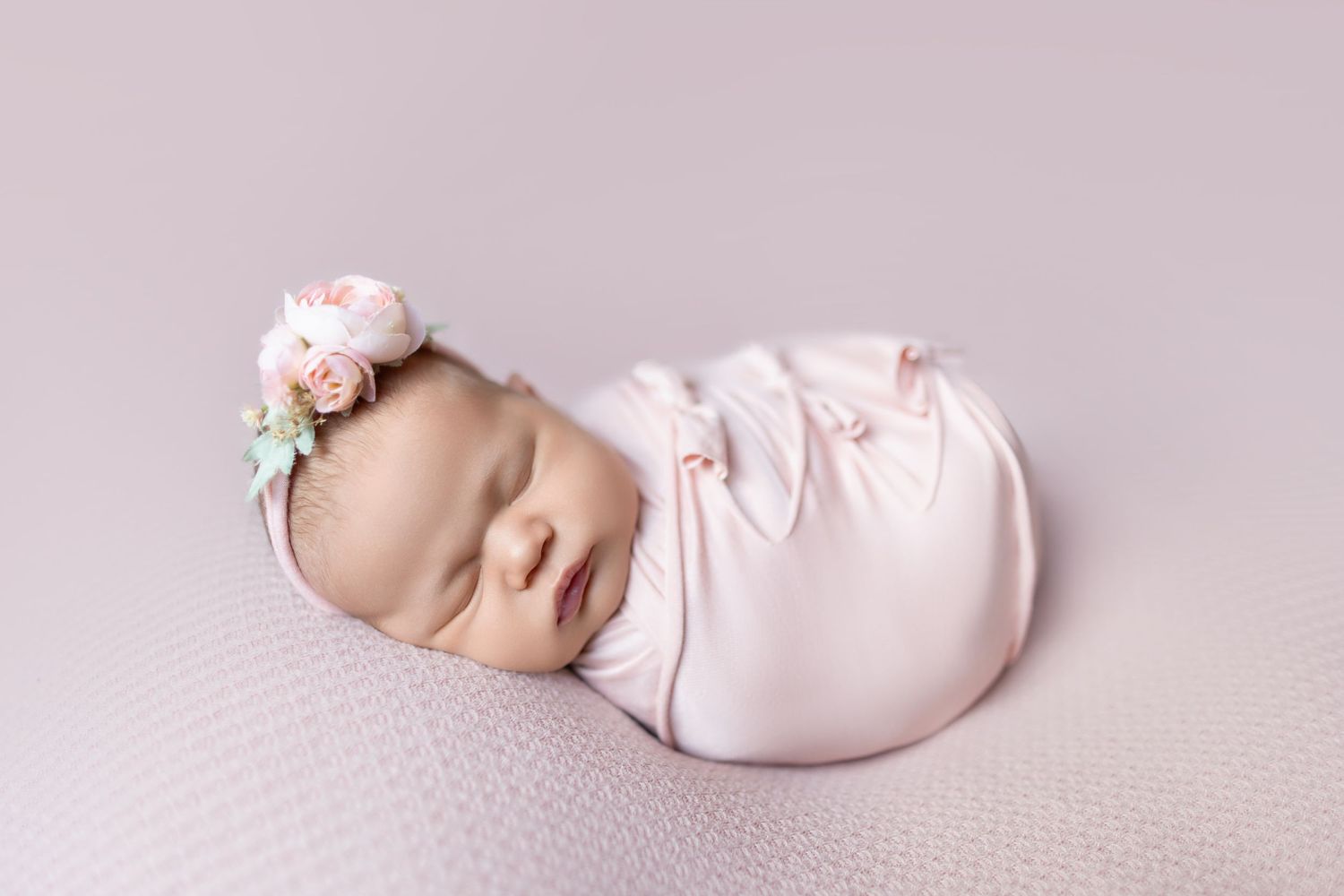 Sleeping newborn baby girl swaddled in pink wrap with floral headband on lavender background.