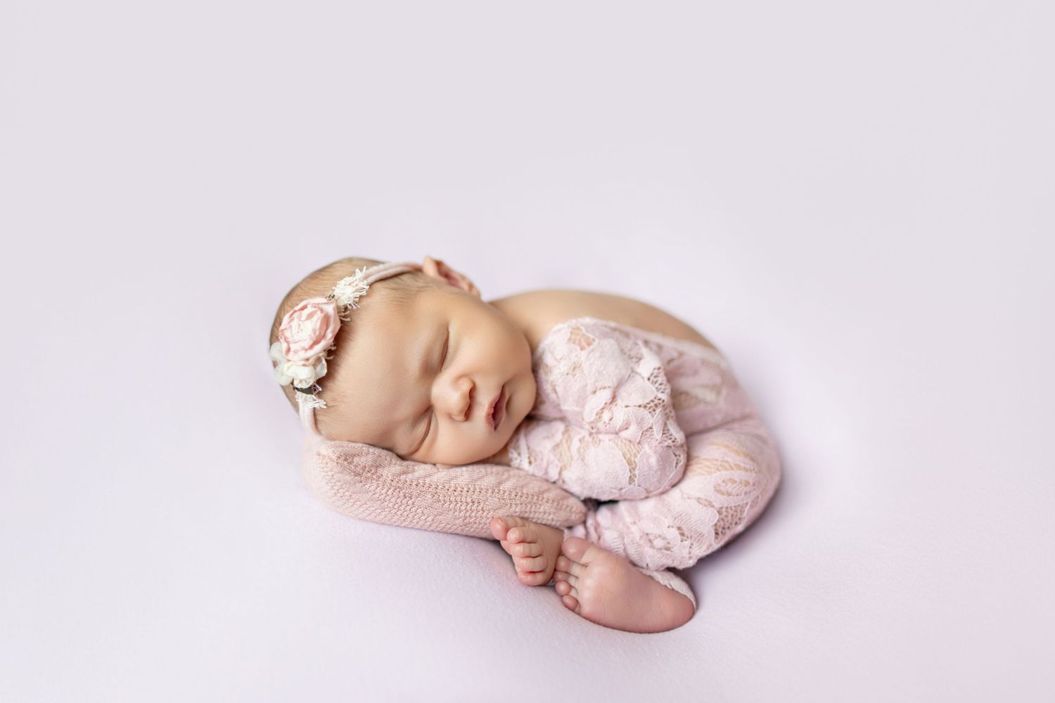 Peaceful newborn baby girl sleeping in pink lace outfit and floral headband on soft white background.