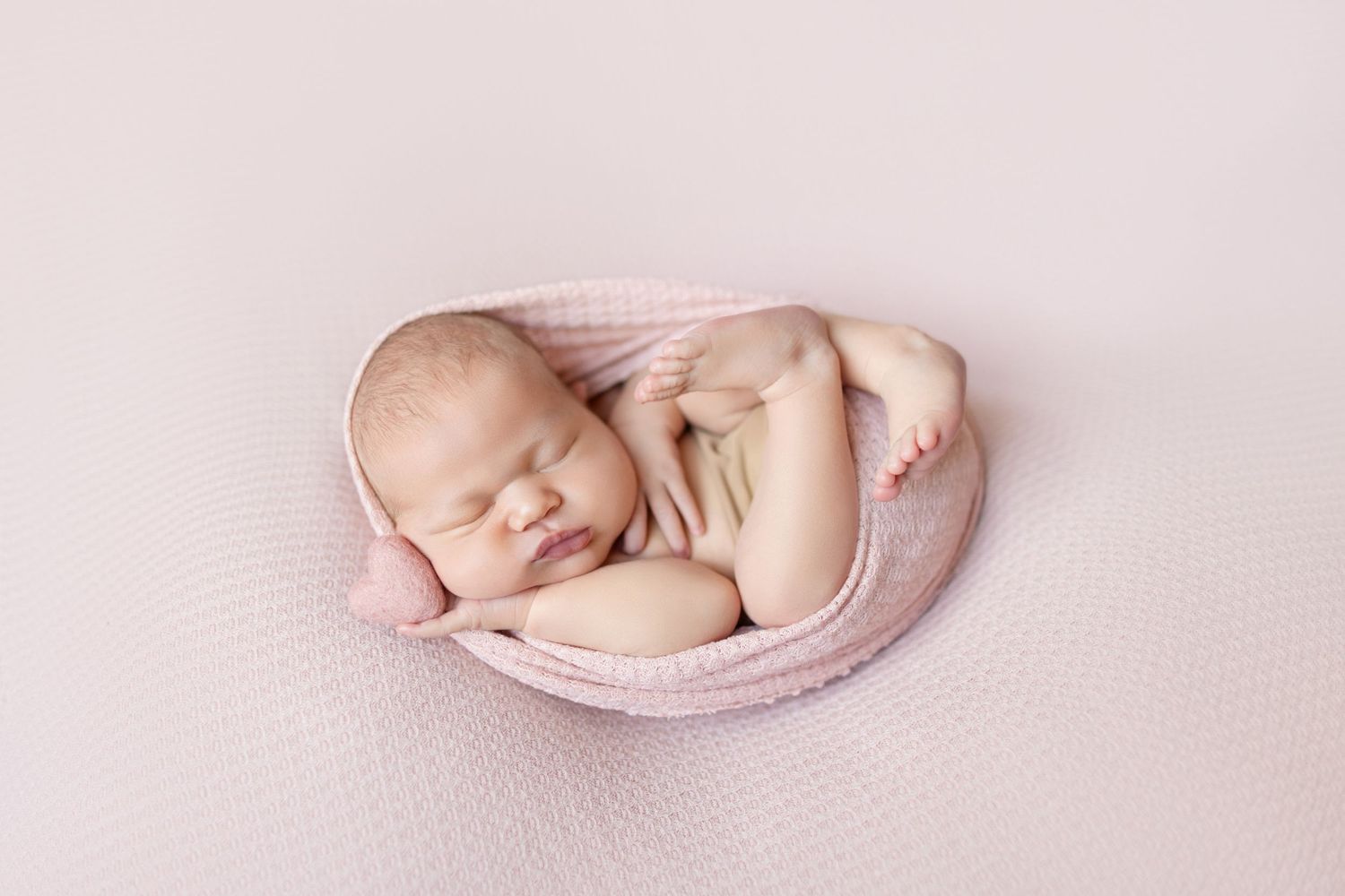 Newborn baby sleeping peacefully wrapped in a soft pink blanket on a pink background.