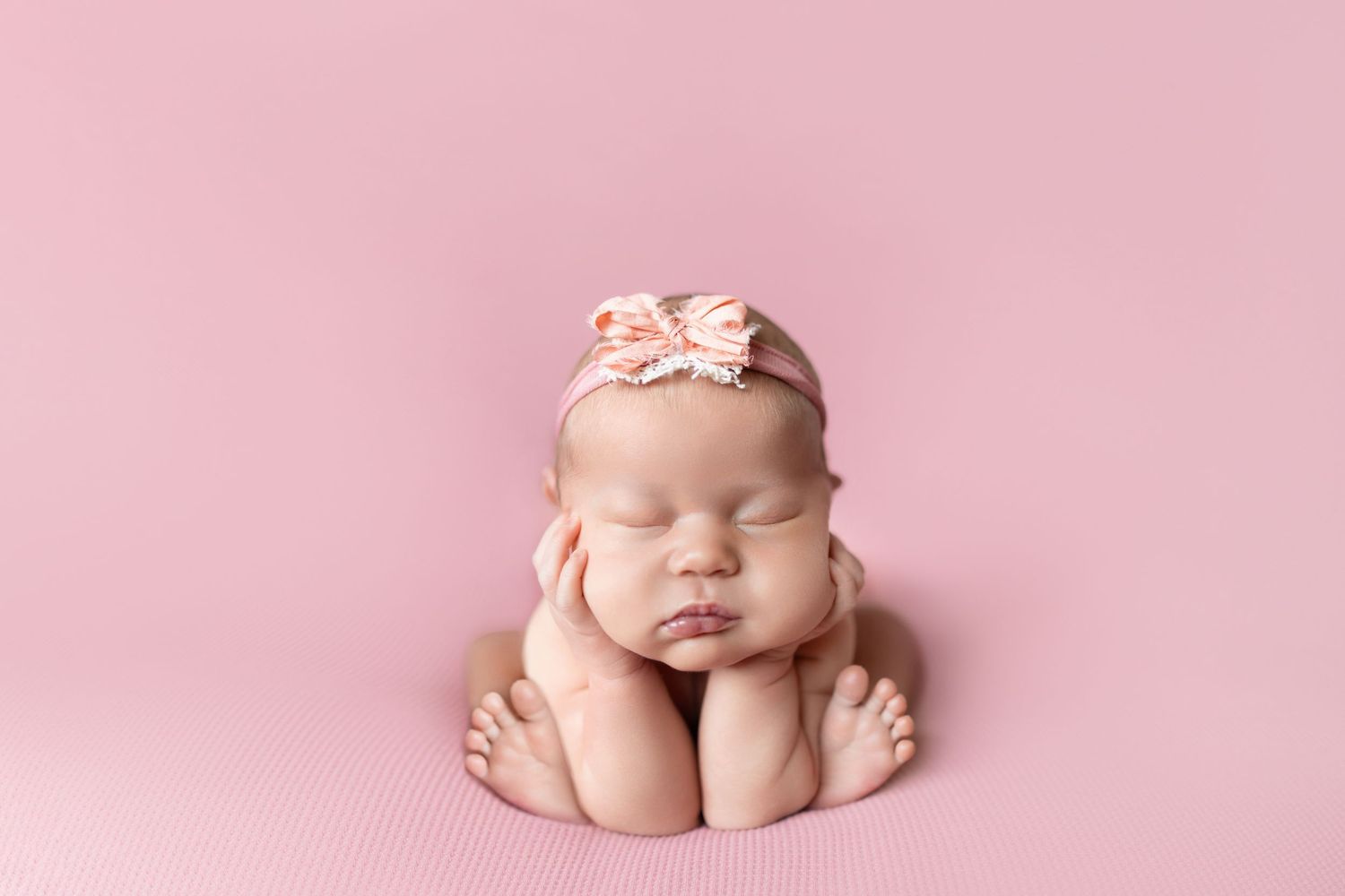 Sleeping newborn baby girl posed on pink backdrop wearing a delicate floral bow headband in a studio portrait.