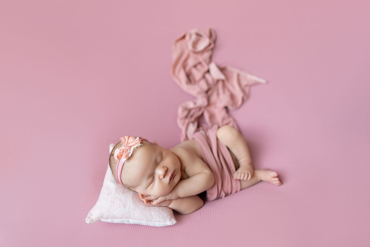 Newborn baby girl sleeping peacefully on pink backdrop, wrapped in pink fabric with floral headband.