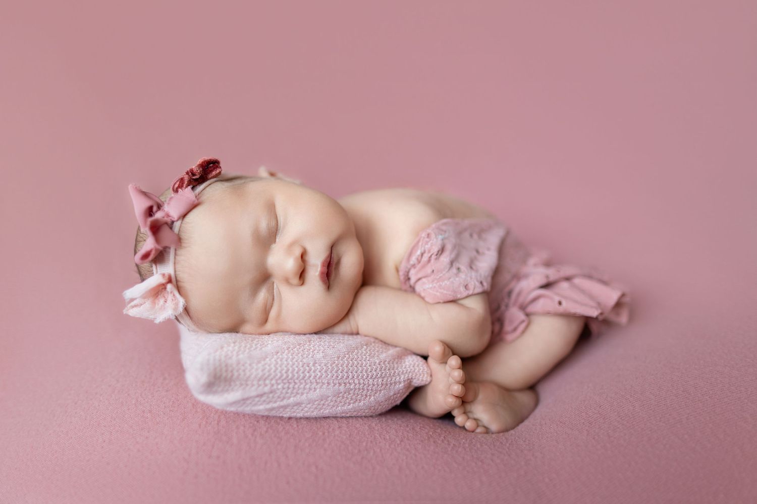 Newborn baby girl sleeping peacefully on pink backdrop wearing pink floral bonnet and ruffled bloomers.