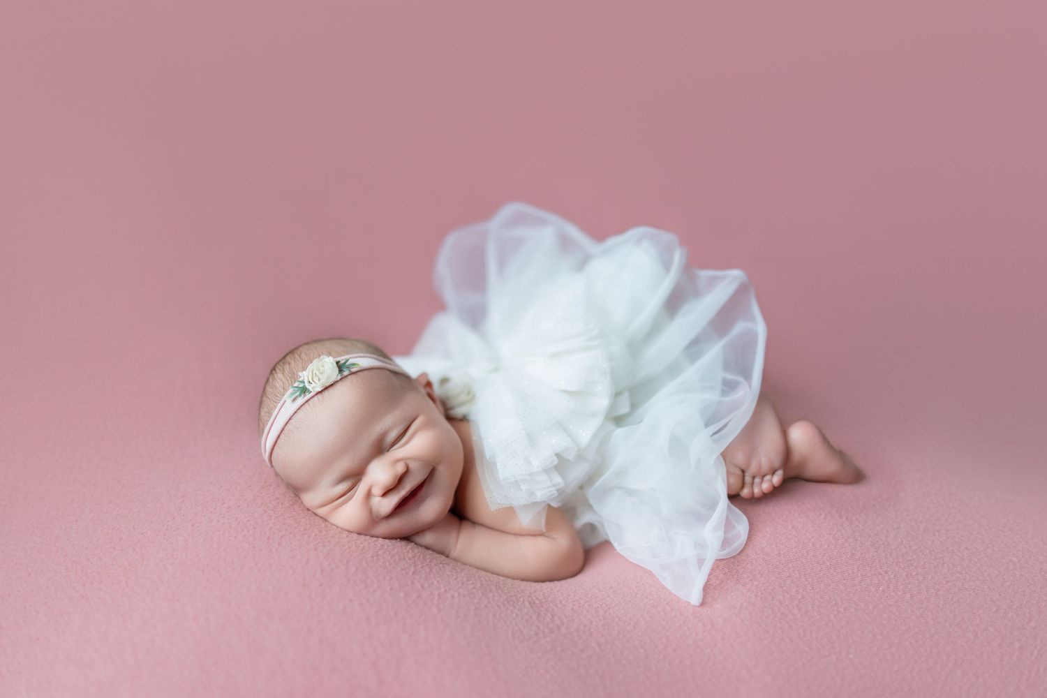 Sleeping newborn baby girl in white tutu dress and floral headband on pink background.