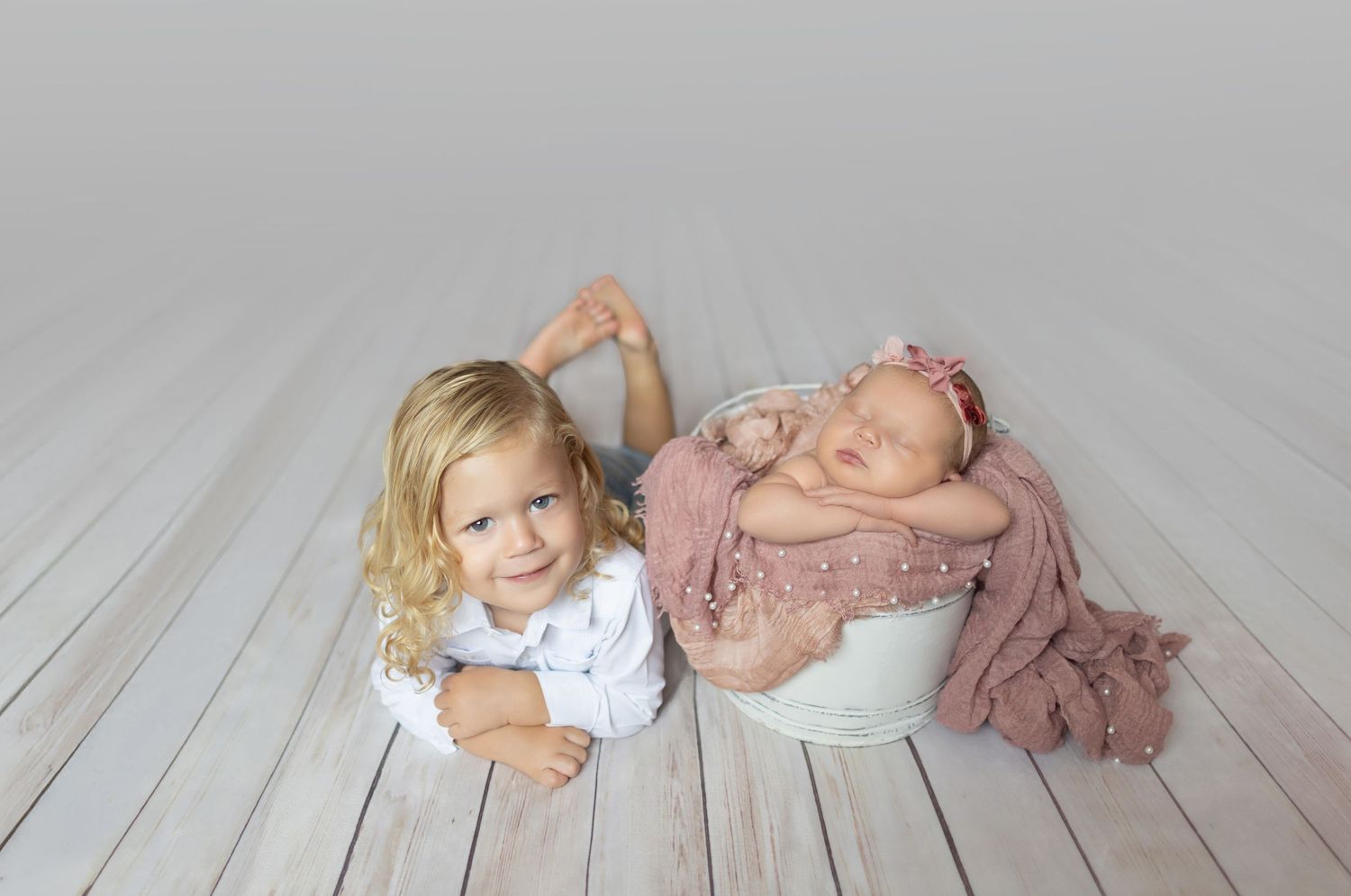 Smiling toddler girl poses beside a sleeping newborn baby wrapped in pink in a white bucket on a wooden floor.