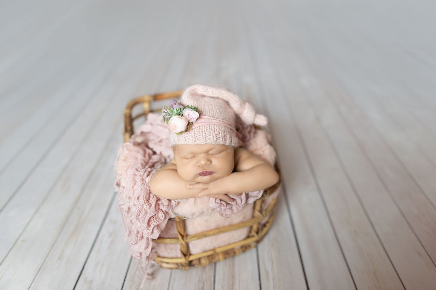 Newborn baby sleeping peacefully in a wicker basket with pink knit hat and floral accents on white wood floor.