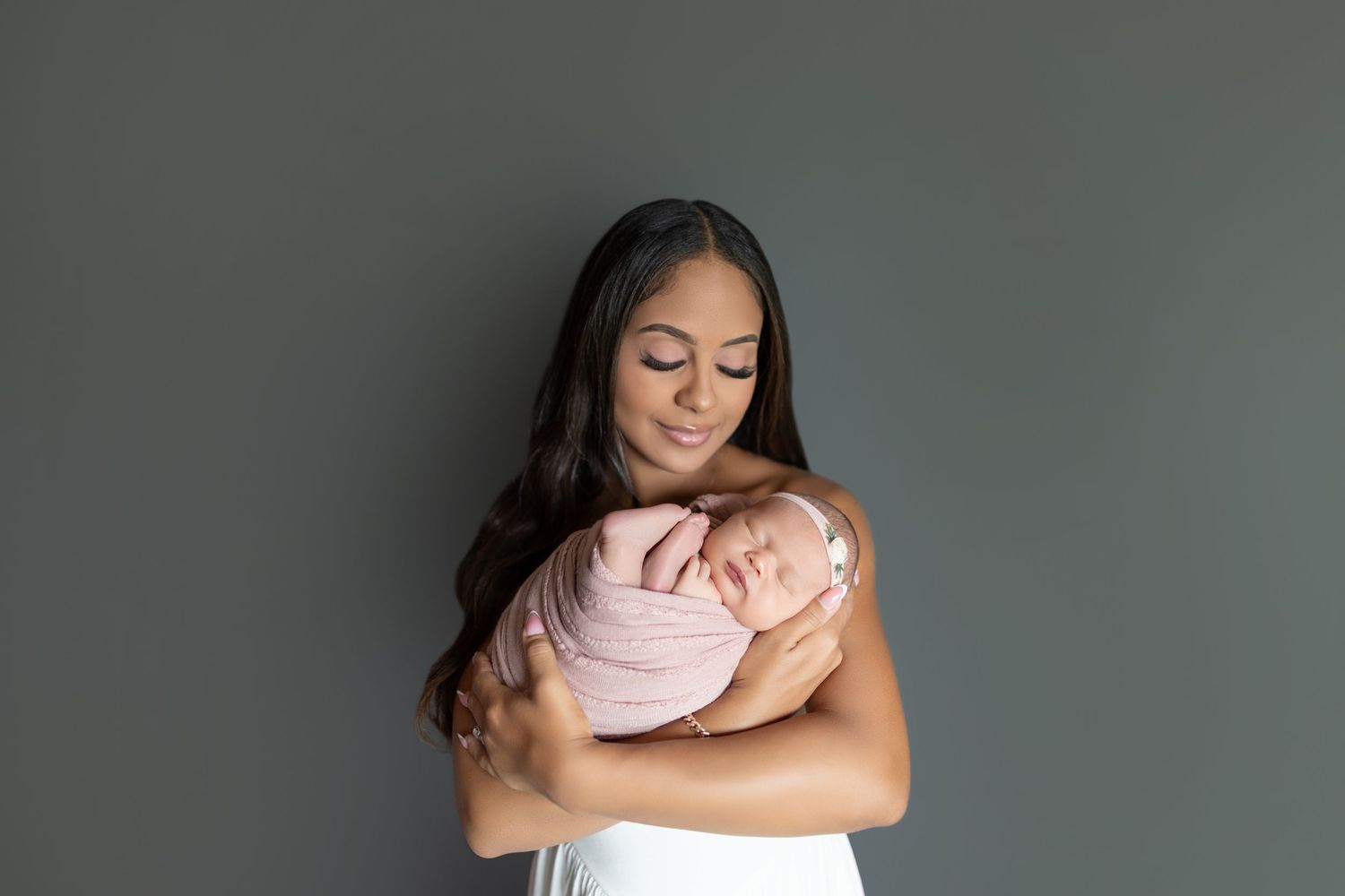 A mother in a white dress tenderly holds her sleeping newborn wrapped in a pink swaddle against a gray background.