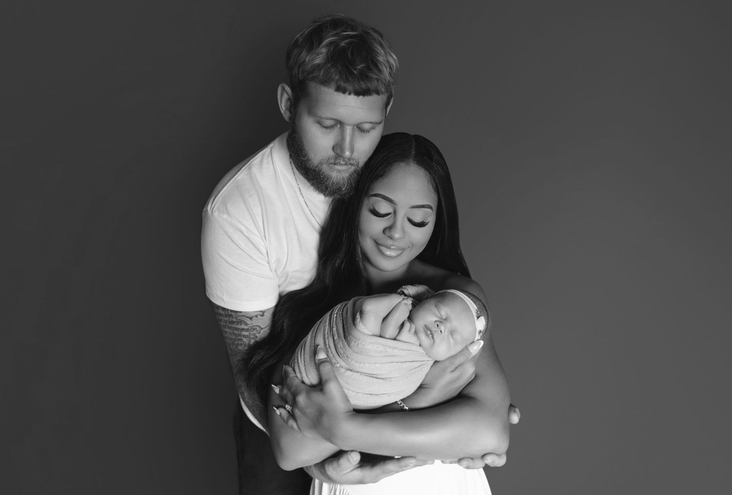 Black and white photo of a couple tenderly embracing their newborn baby in a studio portrait.