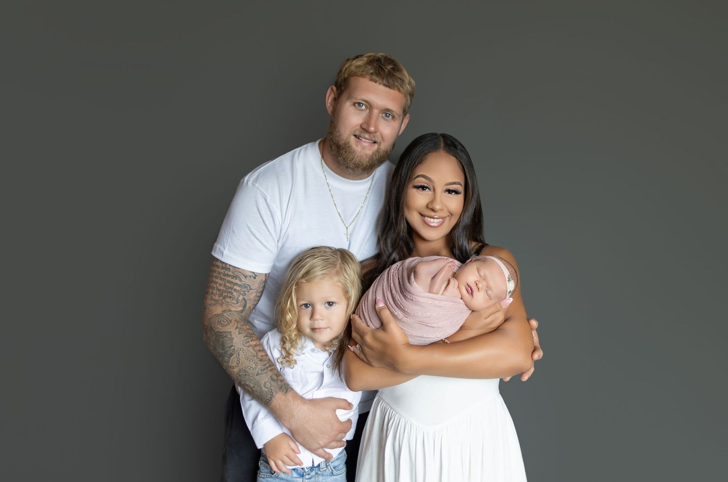 Family portrait of parents with toddler girl and newborn baby posing together against a gray studio background.