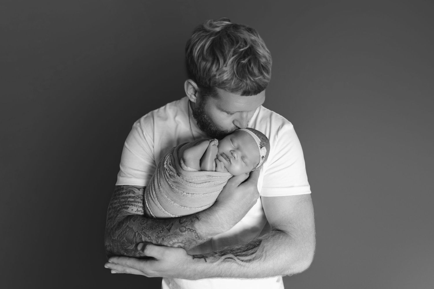 Black and white photo of tattooed father tenderly kissing his swaddled newborn baby.