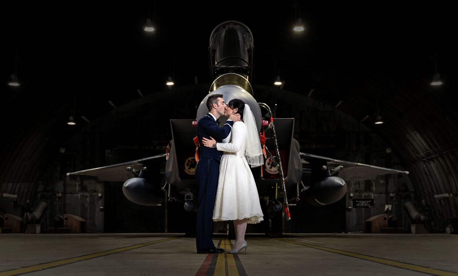 Bride and groom share a romantic kiss in front of a military jet aircraft inside a dark hangar.