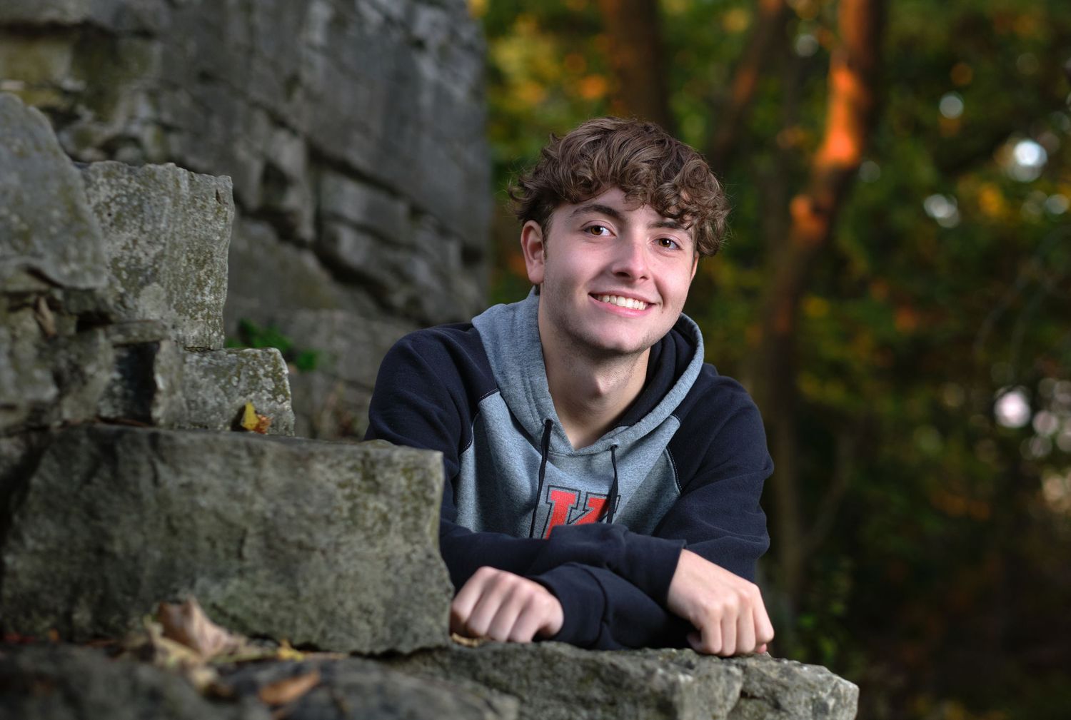 A young man in a hoodie leans on a rocky outcropping while posing for a senior photo in the woods.