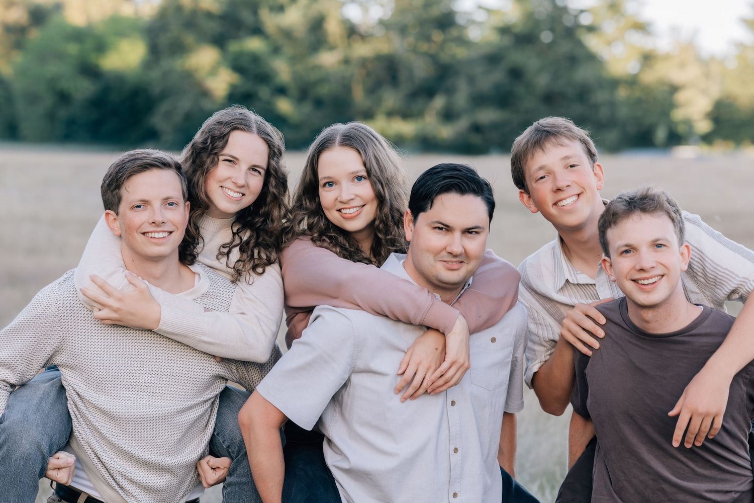 Six smiling young people posing together outdoors in a sunny field for a group family portrait photo.