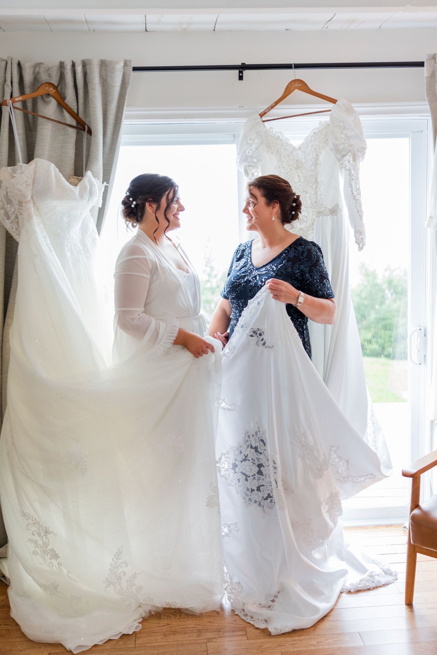 Two women admire a white wedding dress together, surrounded by bridal gowns hanging near a bright window.