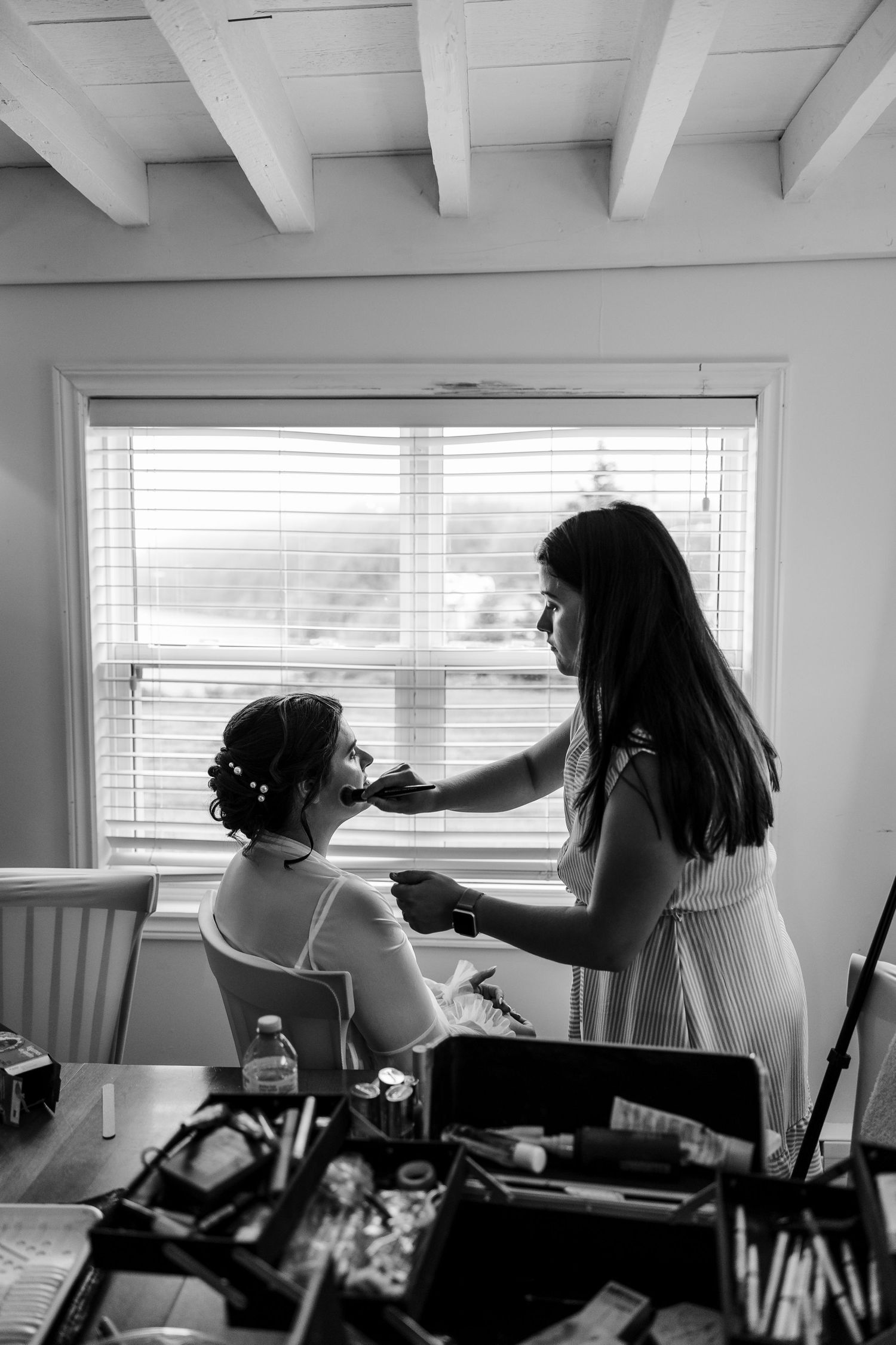 Black and white photo of a makeup artist applying makeup to a bride seated by a window on her wedding day.