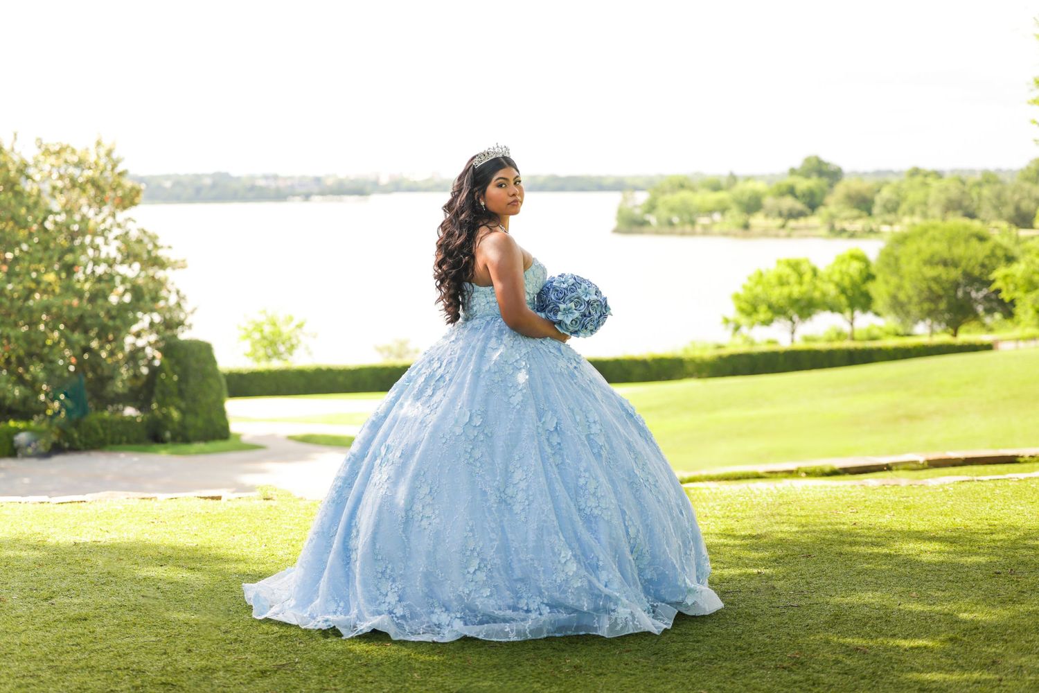 Young woman in a light blue ball gown holding a bouquet, posing outdoors near a lake on a sunny day.
