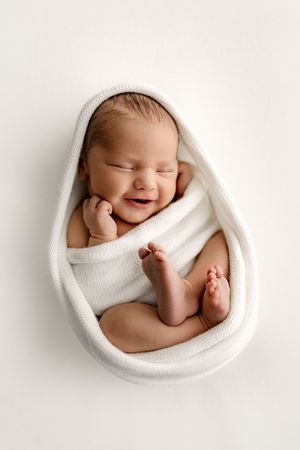 Smiling newborn baby wrapped in white knit blanket, peacefully sleeping with tiny feet visible against white background.