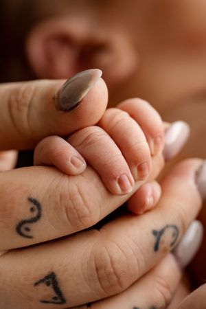 A newborn baby's tiny hand grasping an adult's tattooed finger, with manicured nails visible in soft focus.